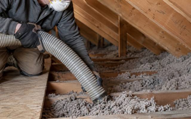 Worker with a vacuum hose taking grey loose-fill out from between the joists during attic insulation removal