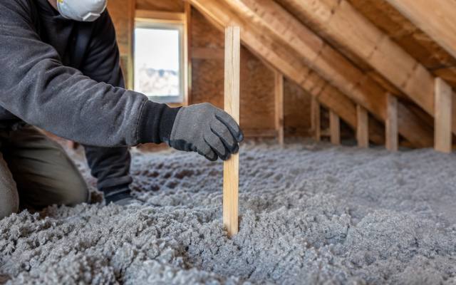 Worker measuring the depth of grey loose-fill insulation with a ruler during an attic check in London
